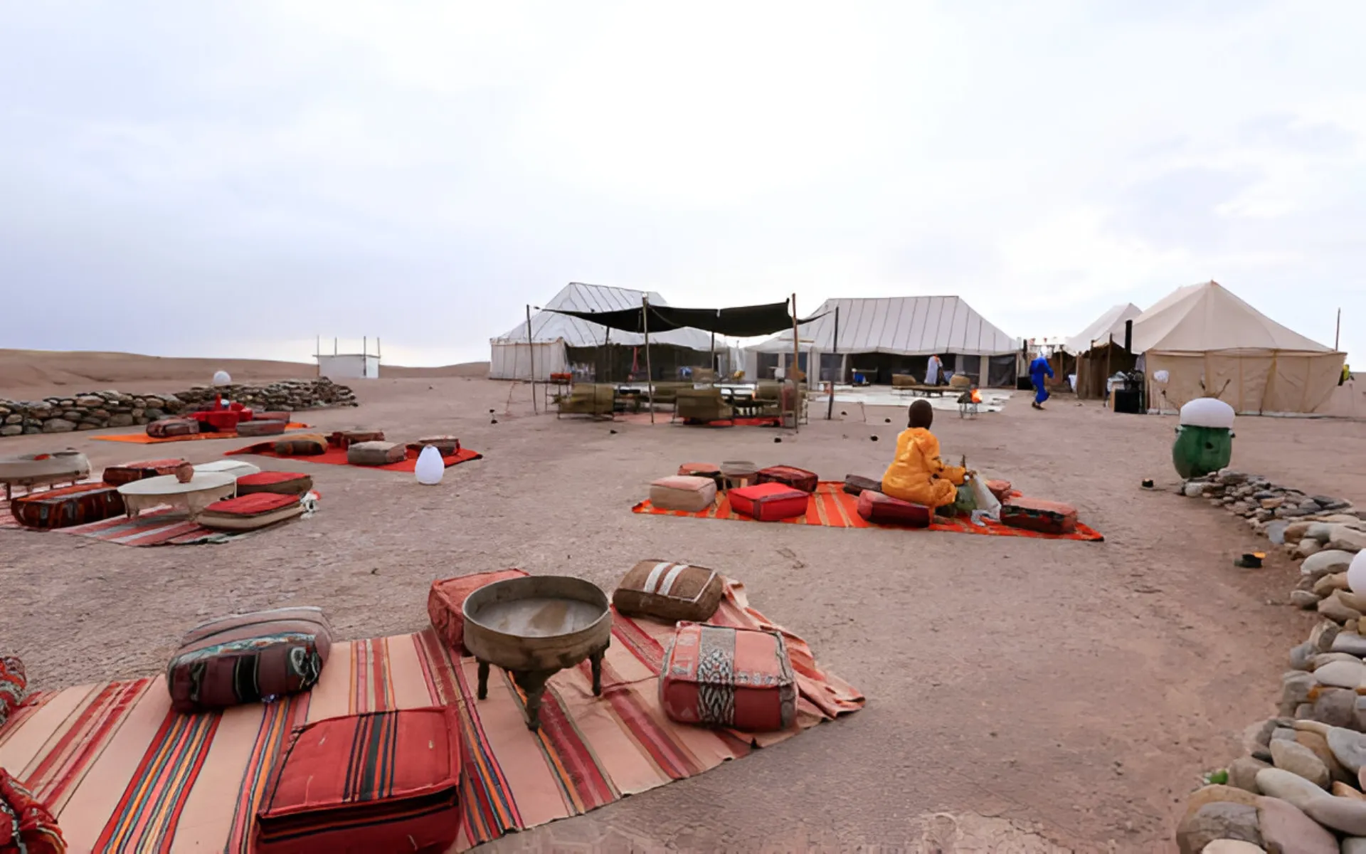Desert camp with striped rugs, low cushions and canvas tents, person seated in yellow