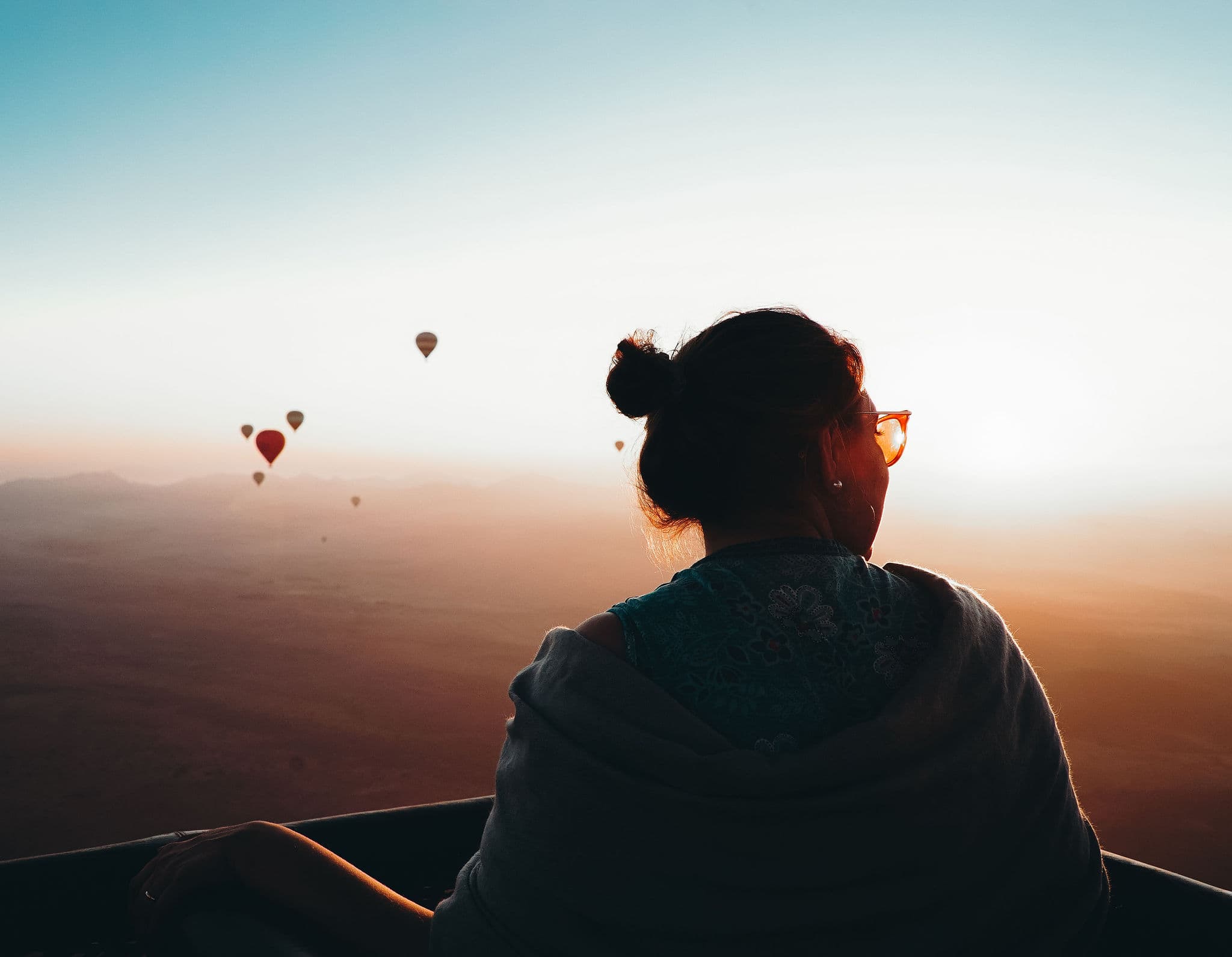 Person silhouetted in balloon basket watching sunrise with distant hot air balloons.